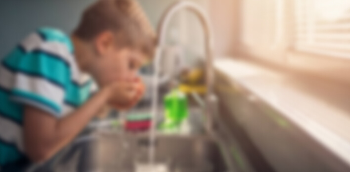 A person leans over a kitchen sink, drinking from hands under a faucet, with colorful kitchen items and sunlight filling the room.