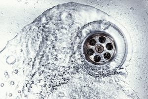 Water drains through a silver sink strainer with small holes, amidst swirling, clear water, forming bubbles in a clean, metallic sink.