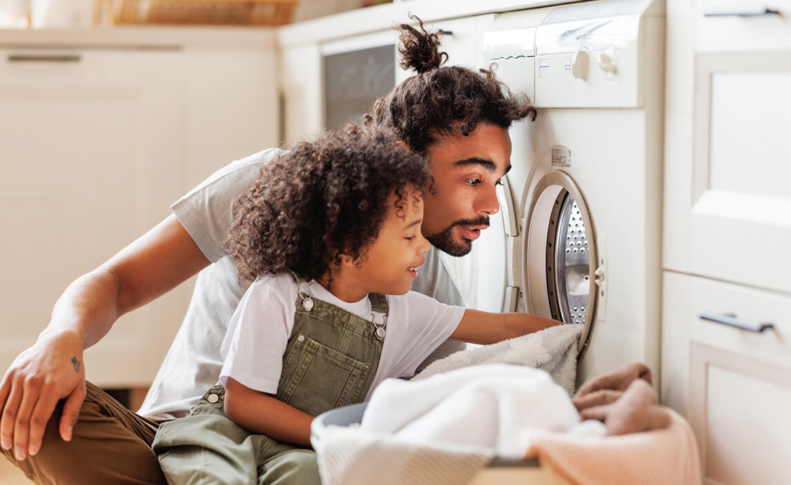 A father and child are crouched beside a washing machine, loading clothes, in a bright kitchen. The child looks curious and engaged.
