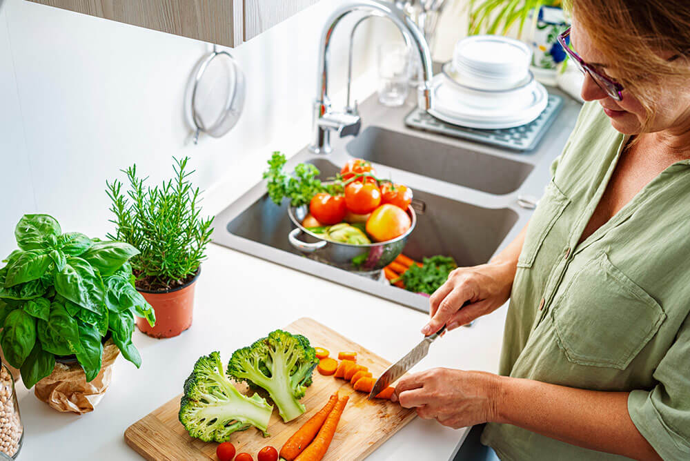 A person chops carrots on a wooden board. Nearby, fresh vegetables and herbs fill a bright kitchen with a sink and clean dishes.