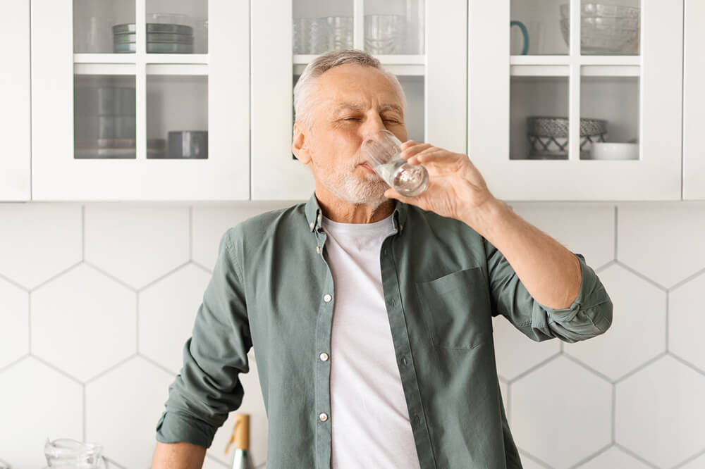 A man drinks from a glass in a bright kitchen with white cabinets and hexagonal tiled walls, appearing relaxed and content.