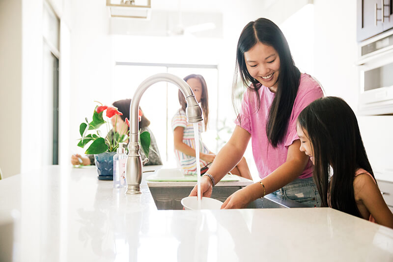 Woman washes dishes with a young girl at a kitchen sink, surrounded by indoor plants and a bright, modern kitchen setting.