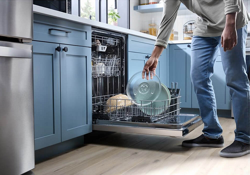 A person loads a dishwasher with plates in a modern kitchen featuring blue cabinets, wooden floors, and various countertop items.